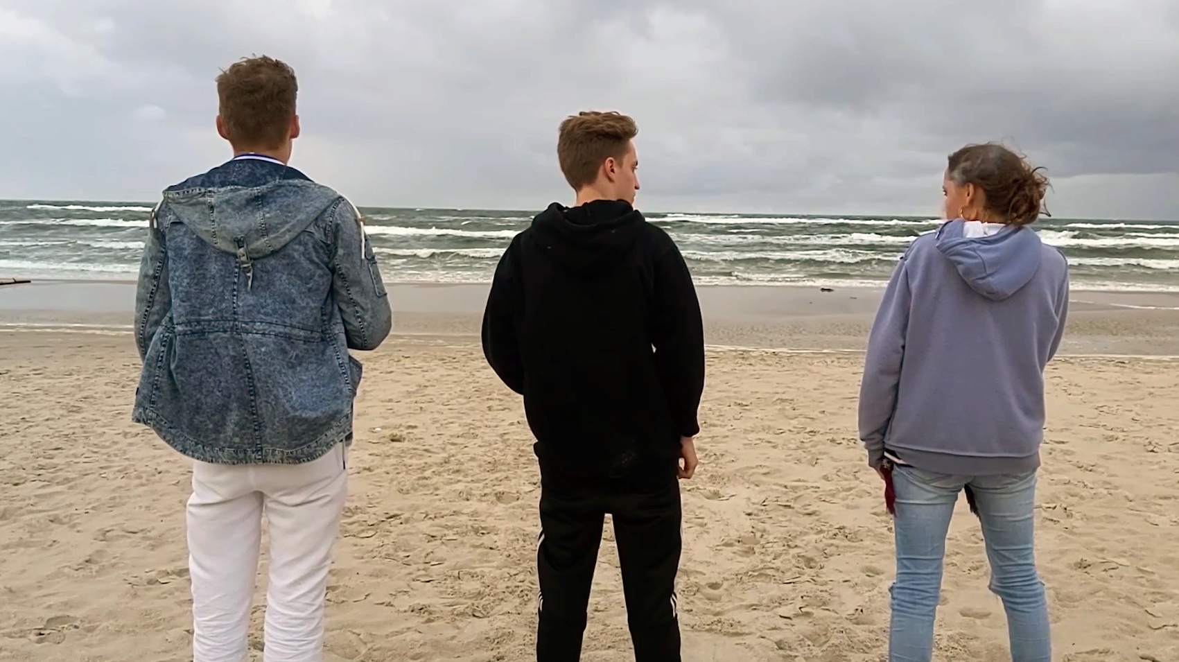 Drei junge Menschen stehen am Strand und schauen aufs Meer.