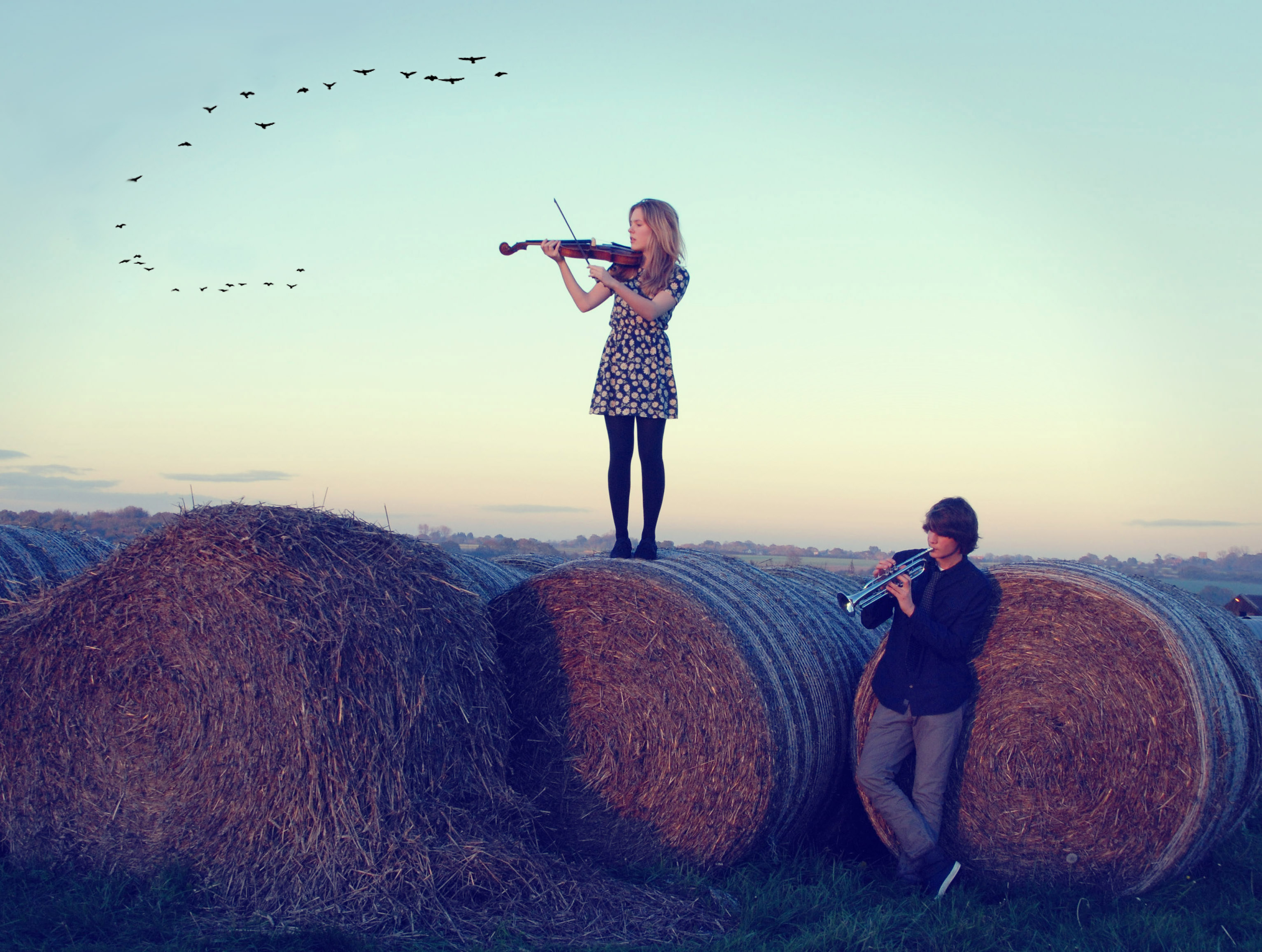 Ein junge Frau steht auf einem von drei Strohballen und spielt Geige. Ein junger Mann vor den Strohballen spielt Trompete. 