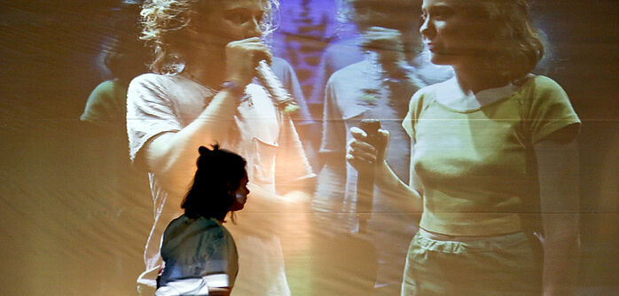 Eine junge Frau vor einer Videoprojektion, die weitere Jugendliche zeigt, die Mikrofone in den Händen halten. 