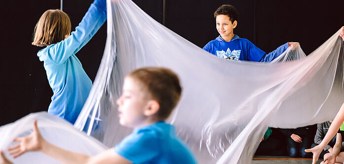 Kinder bewegen ein großes transparentes Tuch auf und ab und lachen dabei.