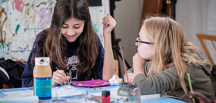 Zu sehen sind zwei Kinder an einem Tisch. Sie halten Pinsel in der Hand und malen, auf dem Tisch stehen Farben und andere Materialien.