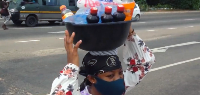 Eine Person mit Mund-Nasen-Schutz trägt eine Schüssel mit Getränkeflaschen auf dem Kopf. 